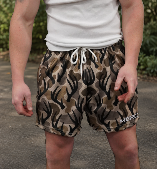 Man wearing MEROX Antler Camo mesh shorts outdoors; approved source-photo logo edit and background adaptation